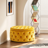 A round yellow tufted ottoman with a matching cushioned lid sits in a bright room with wood flooring, a black-and-white patterned rug, a window with blinds, and a tall magazine rack.
