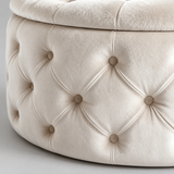Close-up of a tufted round ottoman or stool in a warm cream velvet fabric, showing the diamond button tufting and the soft plush top cushion against a light neutral background.