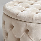 Close-up of a round tufted ottoman or stool in a warm cream-beige velvet fabric, showing the buttoned upholstery and part of the lid against a light neutral background.