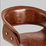 Close-up of the bar stool's curved brown leather backrest, open lower back, stitched edges and gold-tone side studs.
