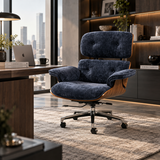 A modern lounge-style office chair with deep blue velvet upholstery and a light wood frame, positioned in a stylish, contemporary home office setting with a desk, laptop, and city skyline view.