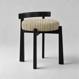 A modern accent chair with a black wooden frame and a round upholstered seat cushion featuring a multicolor geometric pattern on a light beige background, set against a neutral gray studio backdrop.