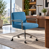 A blue grid-patterned office chair with chrome armrests and a five-wheel base, positioned on a textured rug in a modern, sunlit home office next to a wooden desk.