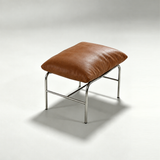 A square ottoman stool with a brown leather cushion on a chrome metal frame, photographed on a light gray studio background.