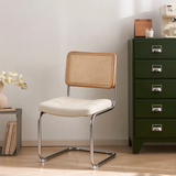 A modern dining chair with a cream upholstered seat, a woven cane backrest, and a chrome metal frame, positioned in a minimalist room next to a dark green cabinet.