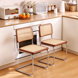 Two modern dining chairs with woven cane seats and backs, brown wooden frames, and chrome metal legs, positioned in a bright, modern kitchen setting.