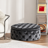 A styled interior scene with a dark gray tufted velvet ottoman in the foreground, a cream lounge chair at left, a tall metal magazine rack with books and boxes at right, and a framed art print on the wall. The product appears to be the ottoman, shown in a real room setting with floor, rug, curtains, and furniture visible.