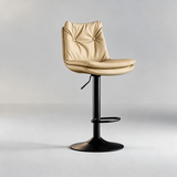 Sand beige bar stool with a button-tufted backrest, padded seat, black pedestal base and oval footrest.