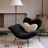 Black ribbed lounge chair with a cream heart cushion beside a floor lamp and large planter in a neutral living room.
