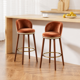 Two rust-brown Alma bar stools with curved backrests and gold-tone footrests beside a light wood breakfast bar.
