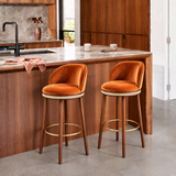 Two orange Alma bar stools with dark wood legs and gold-tone footrests beside a wood-panelled kitchen island.
