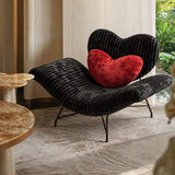 Black ribbed lounge chair with a red heart cushion beside stone-topped tables and sheer curtains near a window.
