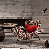 Zebra-print lounge chair with a red heart cushion beside a fireplace and black floor lamp on a striped rug.
