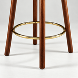 Close-up of the Alma bar stool's dark wood legs and polished gold-tone circular footrest.
