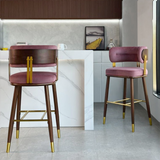 Two modern bar stools with pink velvet seats and backs, dark wood legs, and gold metal accents, shown in a contemporary kitchen setting.