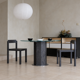 Two black wood dining chairs beside a round glass-top pedestal table beneath a white paper pendant light.
