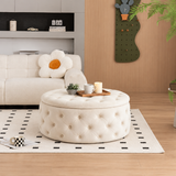 A round tufted ottoman coffee table in a warm cream color, styled in a modern living room setting with a patterned rug, a flower-shaped cushion on a sofa, and decorative accessories on top.