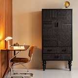 Front view of a tall black carved cabinet with lower drawers beside a desk and brown upholstered chair.