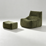 An olive green ribbed fabric lounge chair with a matching square ottoman, photographed on a clean light gray studio floor with soft shadows.