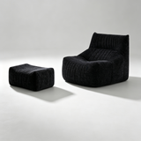 A modern black ribbed velvet lounge chair with a matching square ottoman, photographed on a clean light gray studio floor with dramatic side lighting.